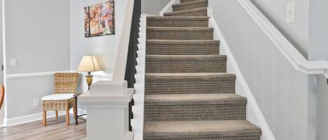 A staircase with a light gray carpet and white railings, leading to a room with a wooden floor and a wicker chair.