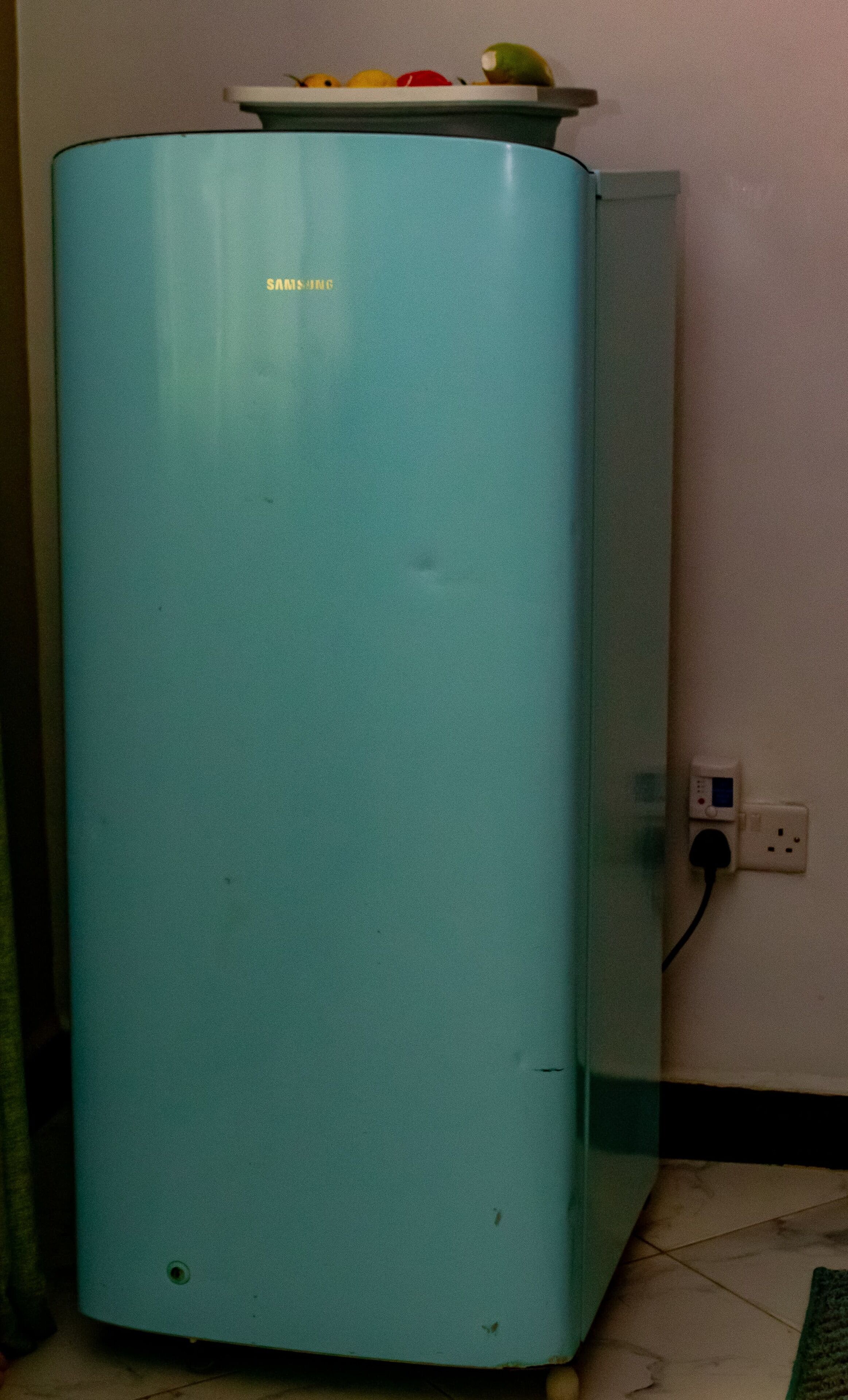 A teal-colored Samsung refrigerator with a fruit bowl on top, positioned next to a wall with a power outlet.