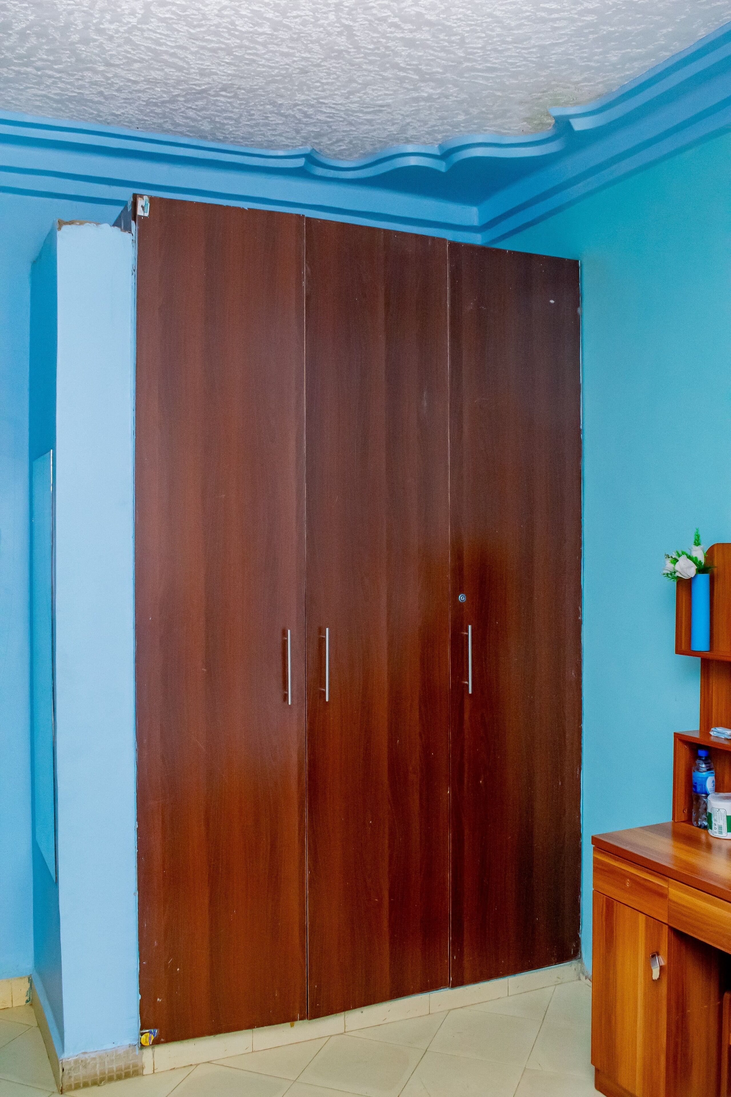 A room with a large wooden wardrobe against a blue wall, a wooden cabinet with a mirror, and a tiled floor.