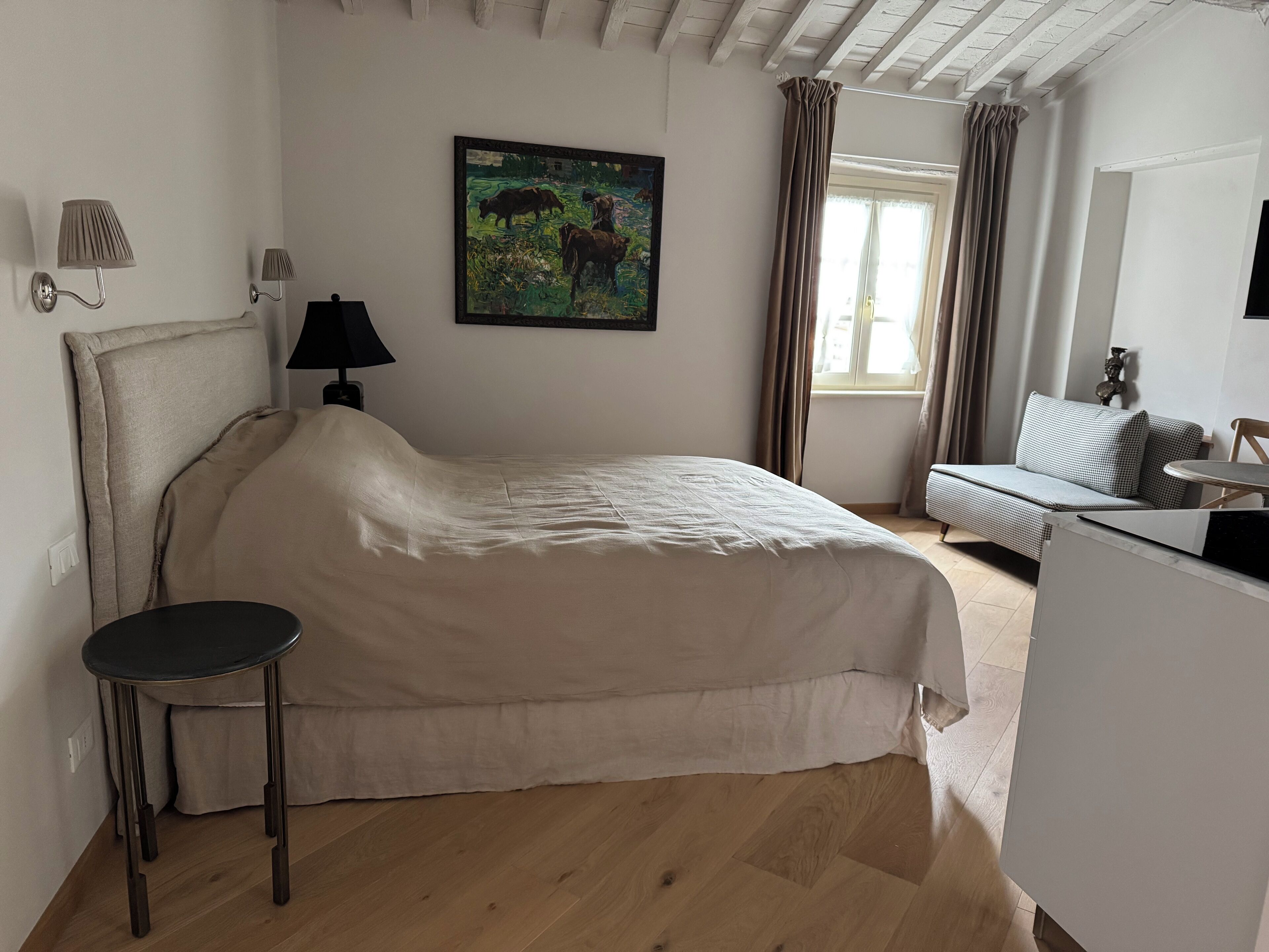 Een slaapkamer met een bed, een klein tafeltje, een bank, een schilderij en een raam met gordijnen.