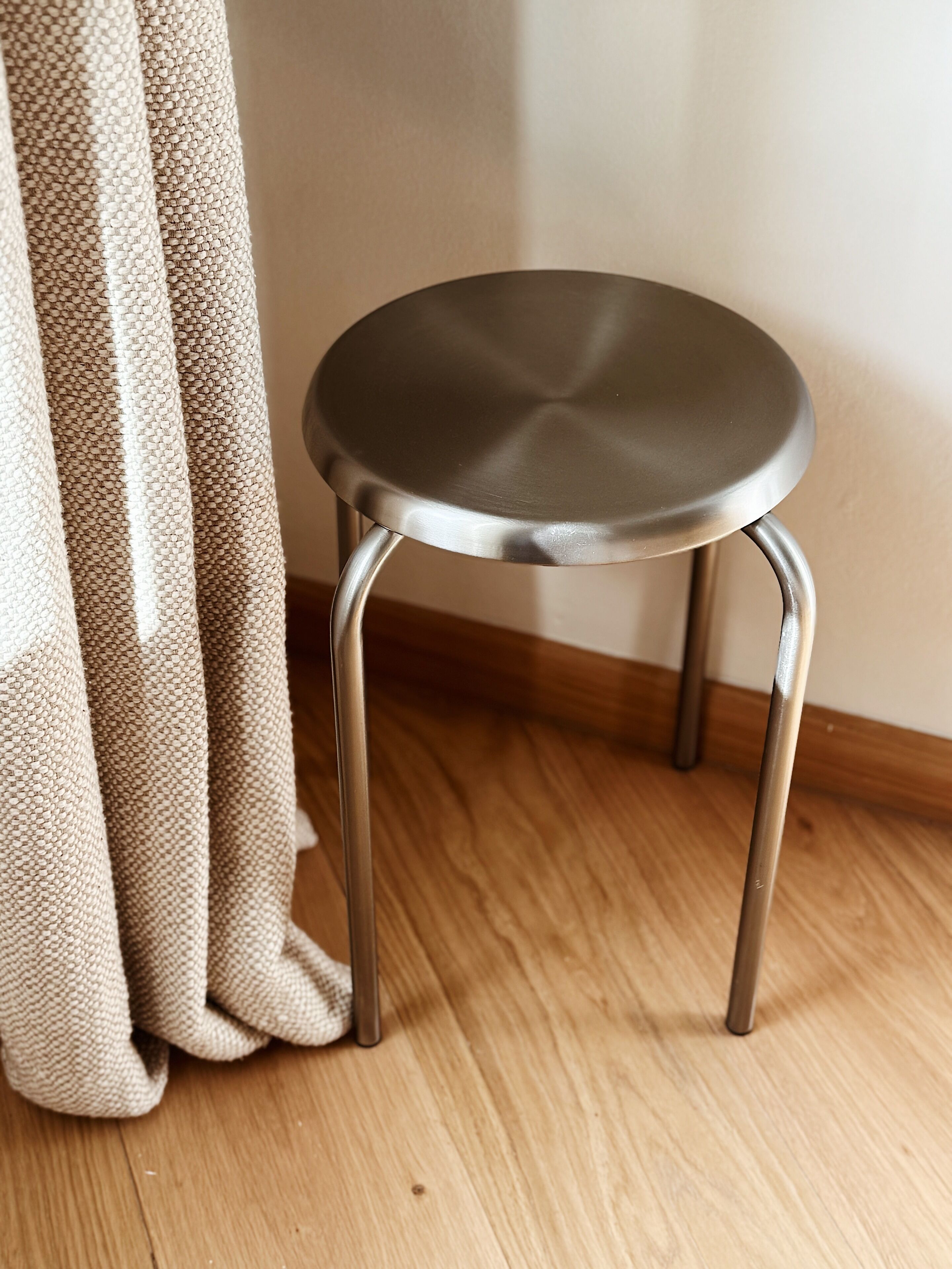 A small, round, metal-framed stool with a smooth surface, positioned on a wooden floor next to a beige curtain.