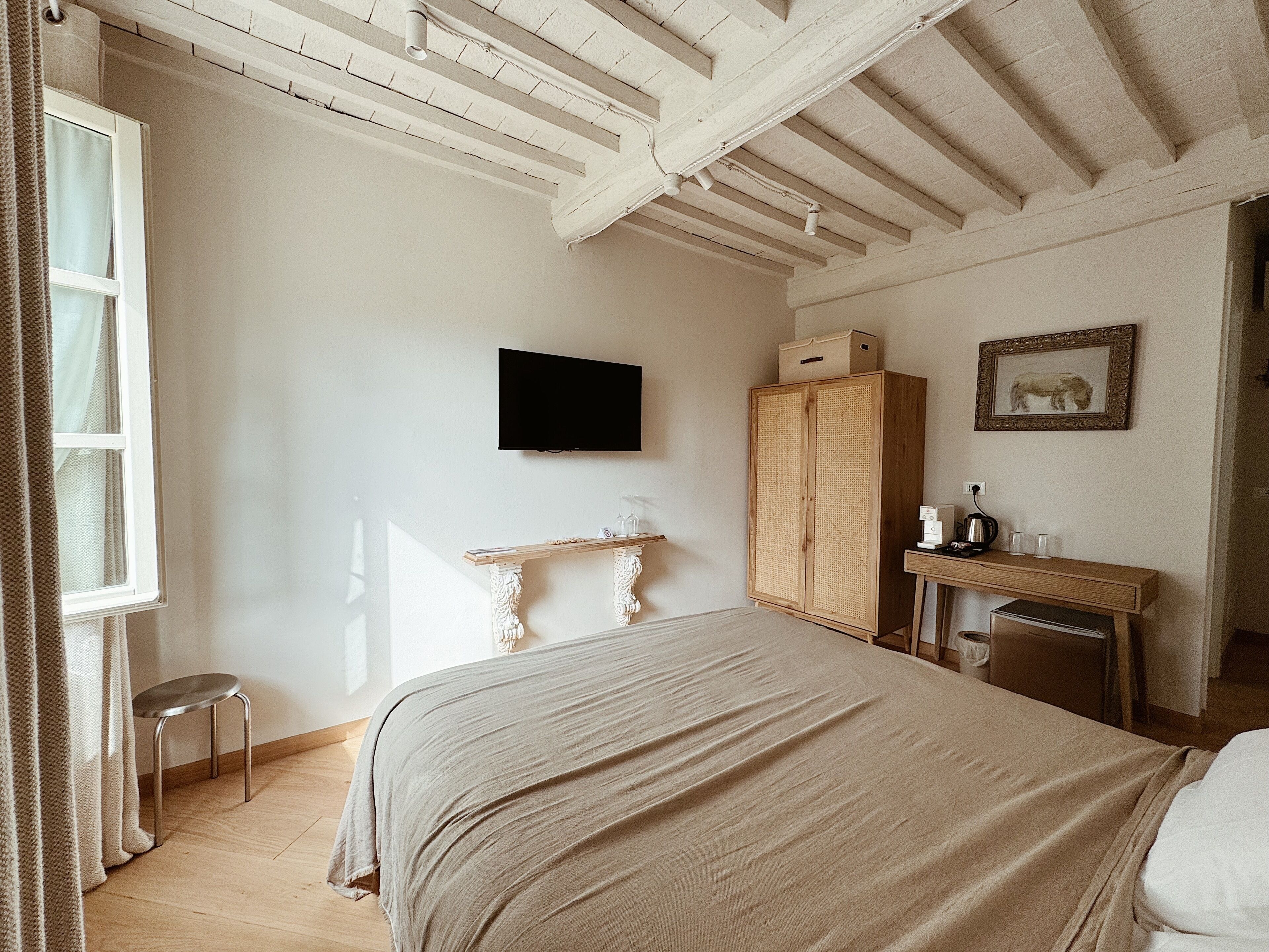 A bedroom with a bed, a television, a wooden wardrobe, a desk with a chair, and a window with curtains.
