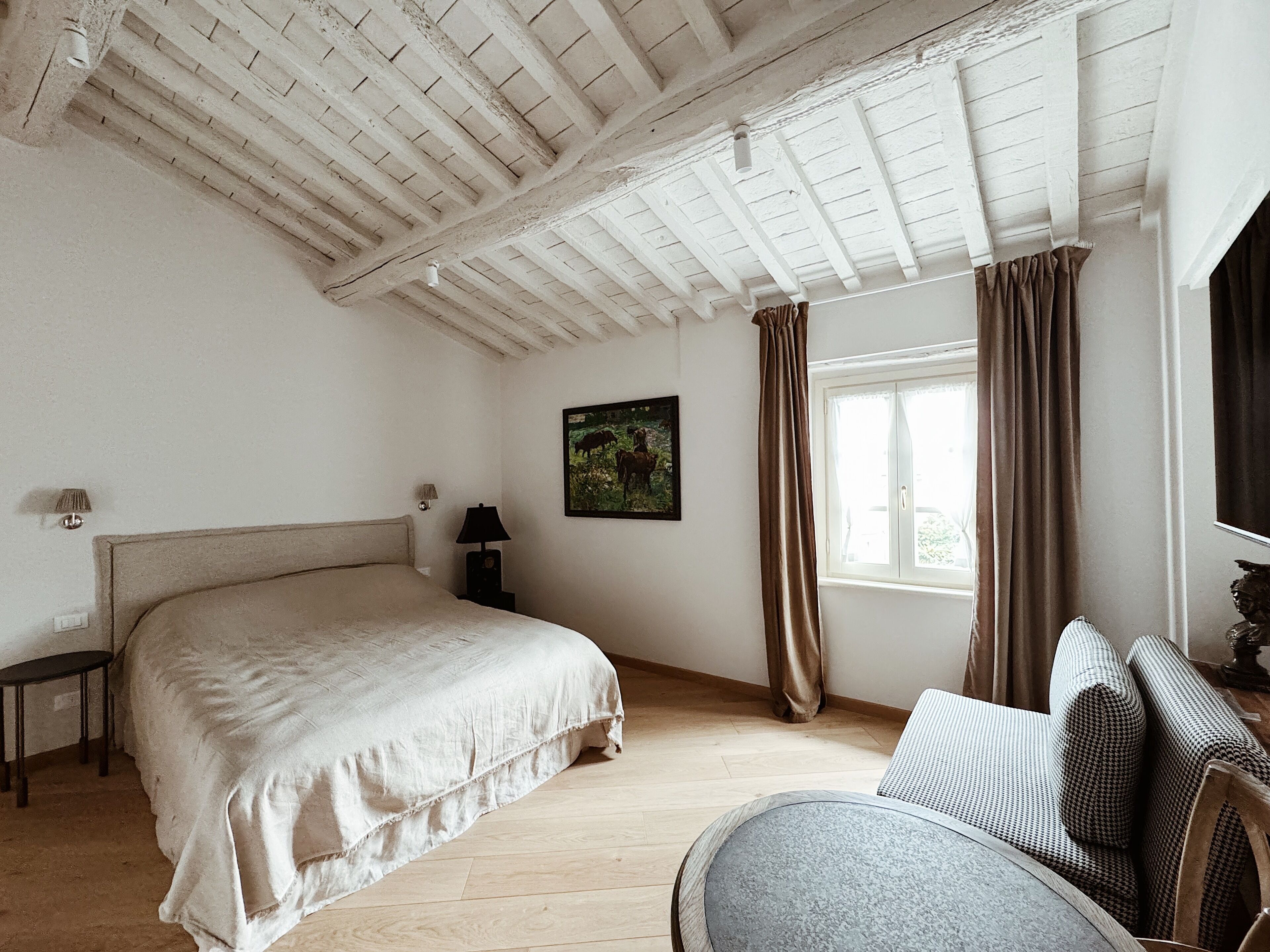 Ein Schlafzimmer mit einem großen Bett, einem Nachttisch, einer Lampe, einem Fenster mit Vorhängen und einem Bild an der Wand.