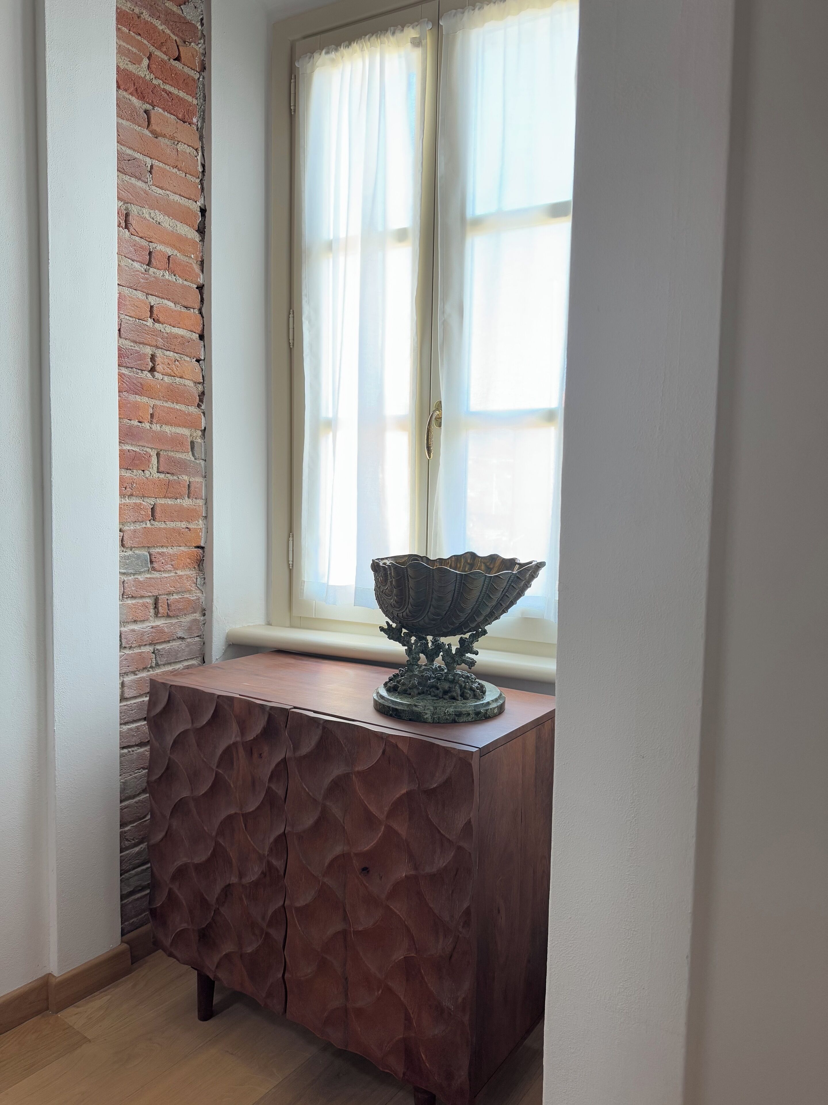 Una habitación de esquina con pared de ladrillo, un mueble de madera con diseño texturizado y una ventana con cortinas translúcidas.