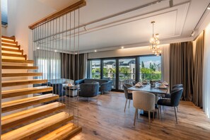 Ein modernes Interieur mit einer Holztreppe, einem Essbereich inklusive Tisch und Stühlen sowie großen Fenstern mit Blick ins Freie.
