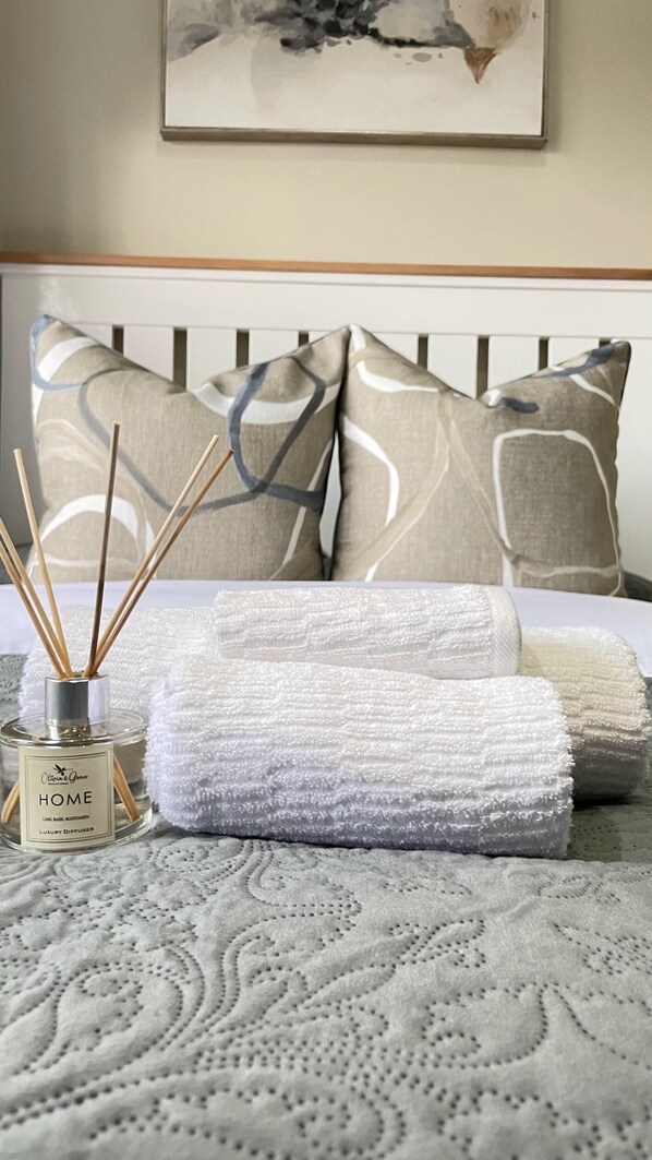 A neatly made bed with patterned pillows, a bottle of home fragrance, and rolled towels on a textured bedspread.