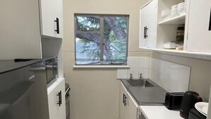 A compact kitchen with white cabinets, a built-in sink, and a window with blinds. - The Comfy Private Place - 2296 (Harare)