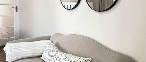A neutral-colored sofa with a white and black polka dot pillow, two wall-mounted mirrors, and a floral painting.