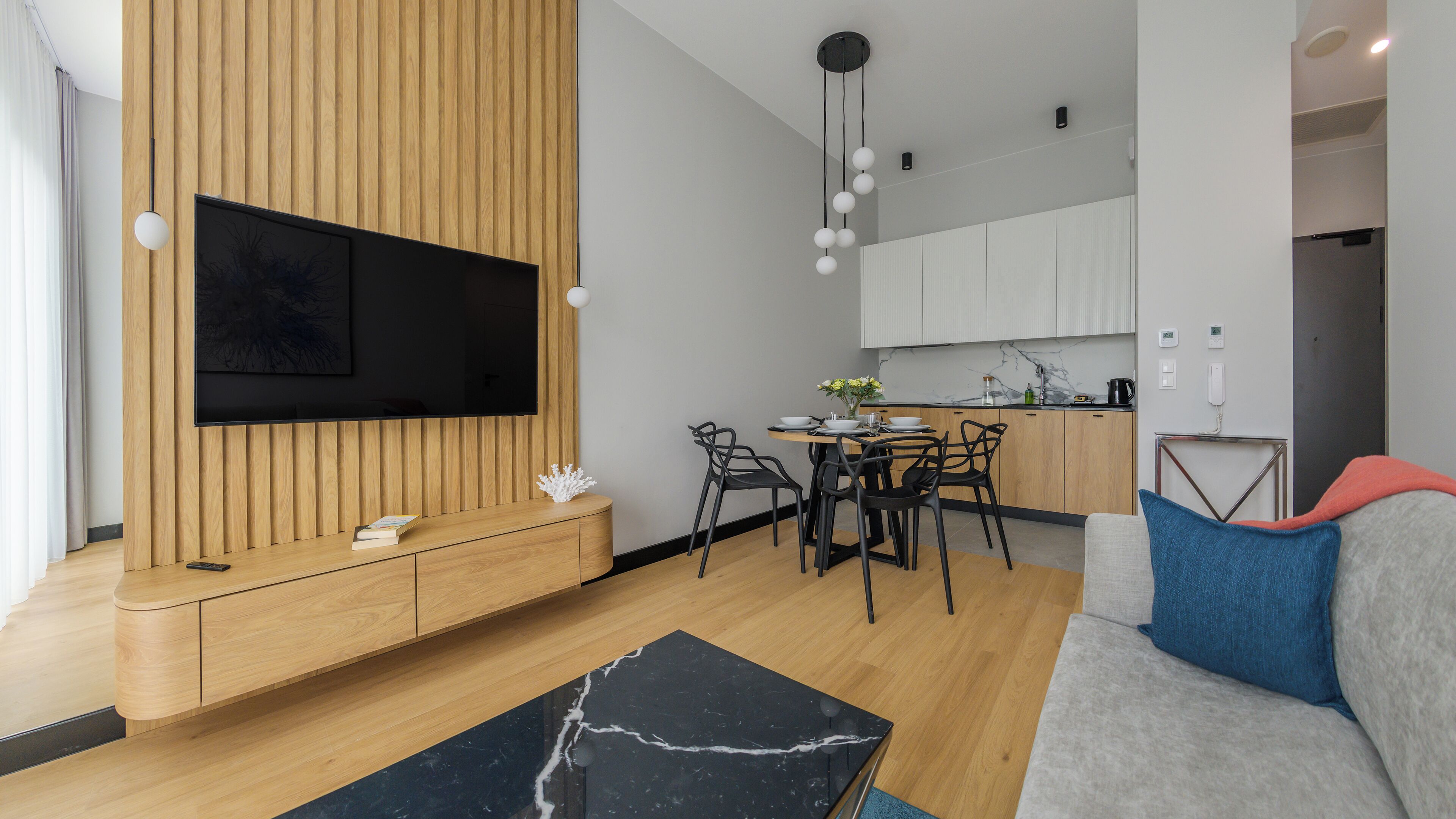 A modern living room with a flat-screen TV, wooden panel wall, black and white marble coffee table, and a grey sofa with blue cushions.