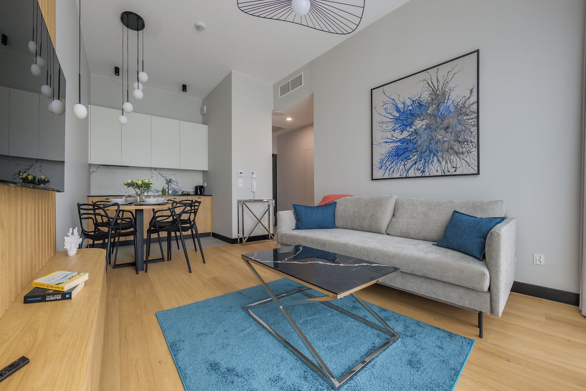 A modern living room with a grey sofa, blue cushions, a coffee table, and a blue rug. There is a dining area with a wooden table and chairs, and a kitchen in the background.
