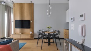 Una sala de estar moderna con un televisor de pantalla plana montado en una pared de paneles de madera, una zona de comedor con una mesa redonda y sillas negras, y una cocina con encimera negra y electrodomésticos blancos.