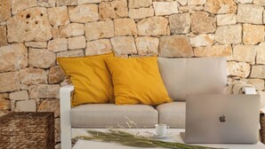 Una sala de estar moderna con una pared de piedra, un sofá con cojines amarillos, una mesa de centro blanca con una computadora portátil, una taza y pastos decorativos.