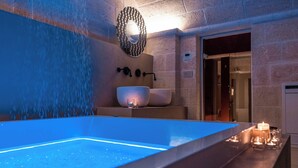 Un baño moderno con una bañera grande iluminada, un espejo fijado a la pared y una mesita de noche con velas.