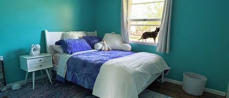 A bedroom with teal walls, a bed with blue and white bedding, a window with a view of the outdoors, and a white rug.