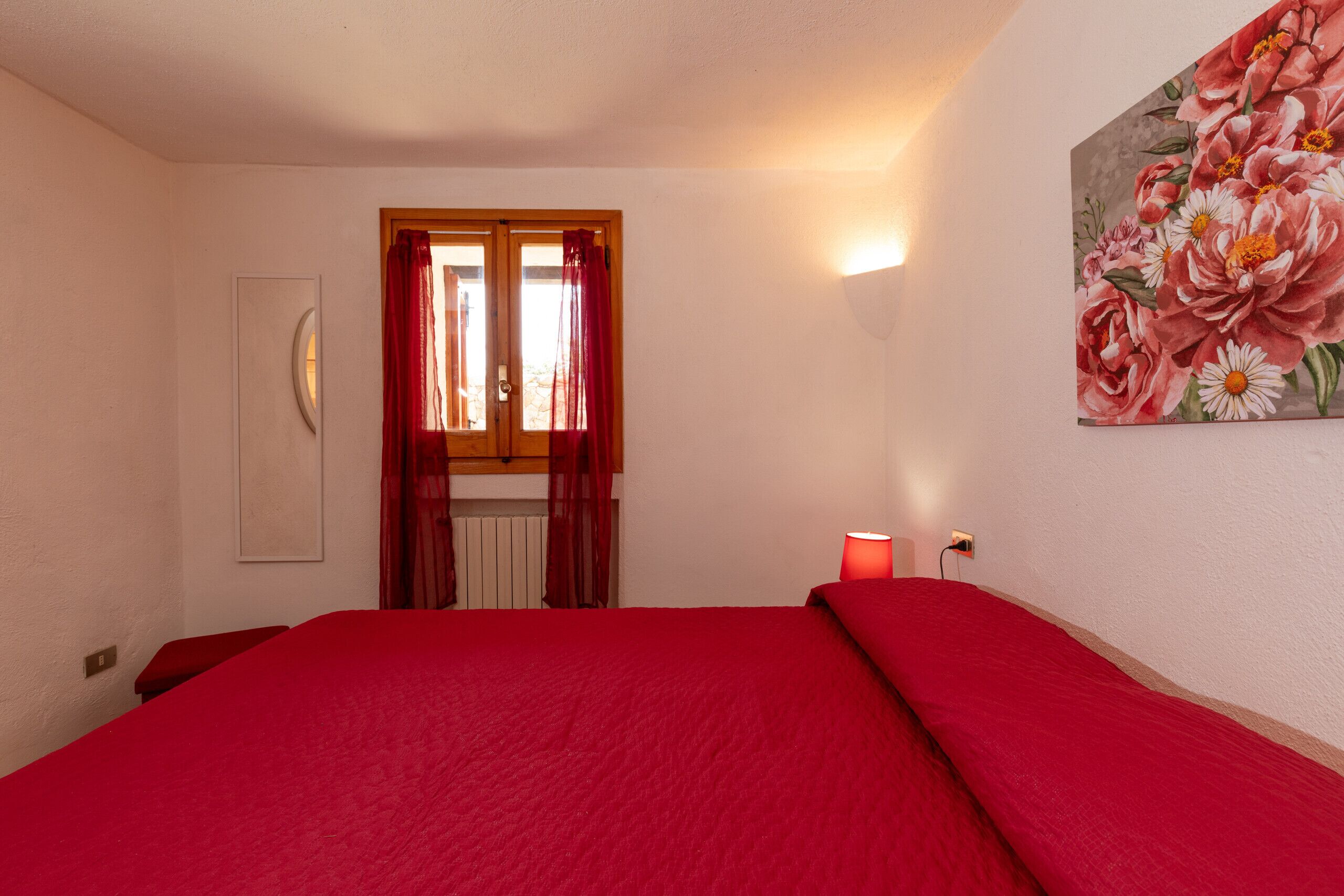 Een slaapkamer met een rood beddegoed, een bloemen schilderij aan de muur, en een raam met rode gordijnen.
