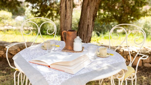A round table with a lace tablecloth, surrounded by white metal chairs, set outdoors with a tree in the background. - Casa Abba by Wonderful Italy (Golfo Aranci)