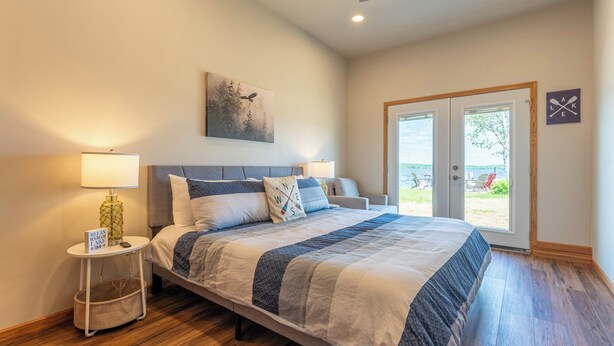 A bedroom with a bed, two bedside tables, a ceiling fan, a painting, and a door leading to a balcony with outdoor seating.