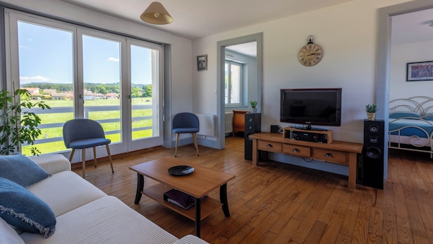 Una sala de estar moderna con sofá, sillas, una mesa de centro, televisor y un altavoz. Amplias ventanas ofrecen vistas a un paisaje verde.
