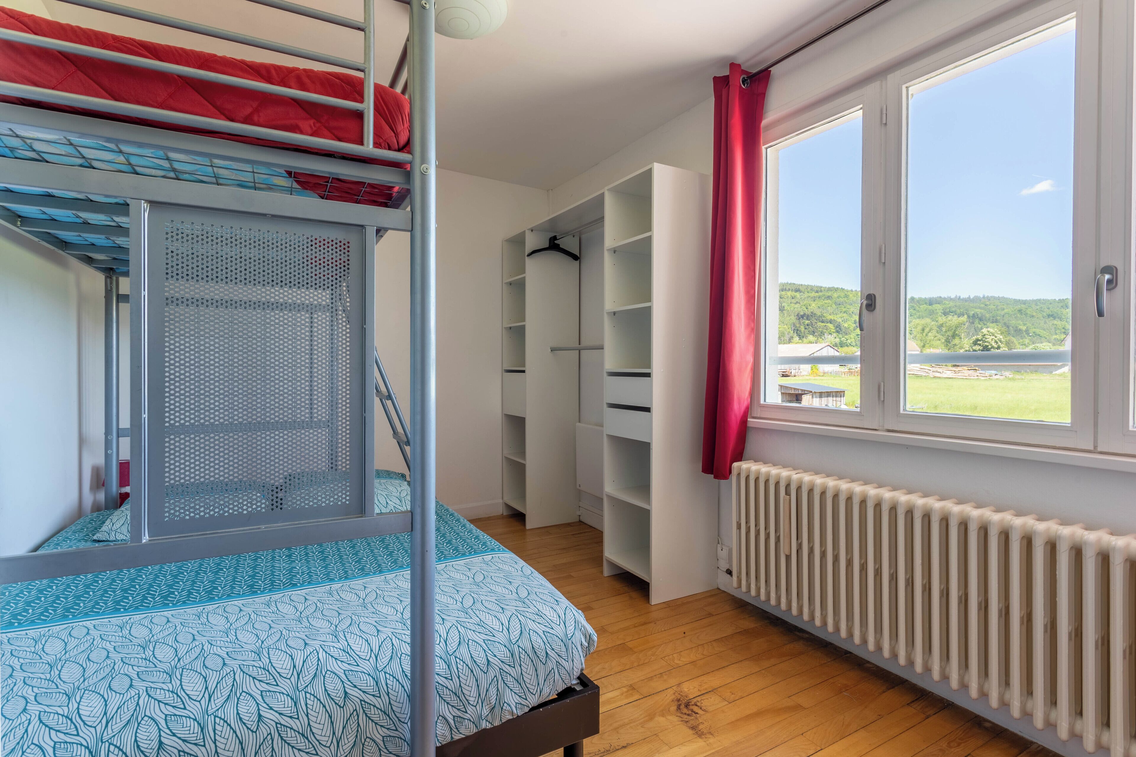 Een stapelbed met een bedspreider met patroon, een inloopkast met planken en uitzicht op een groen landschap door een raam.