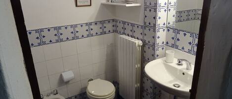 A small bathroom with a toilet, bidet, and sink. There is a framed certificate on the wall and a shelf with toiletries.