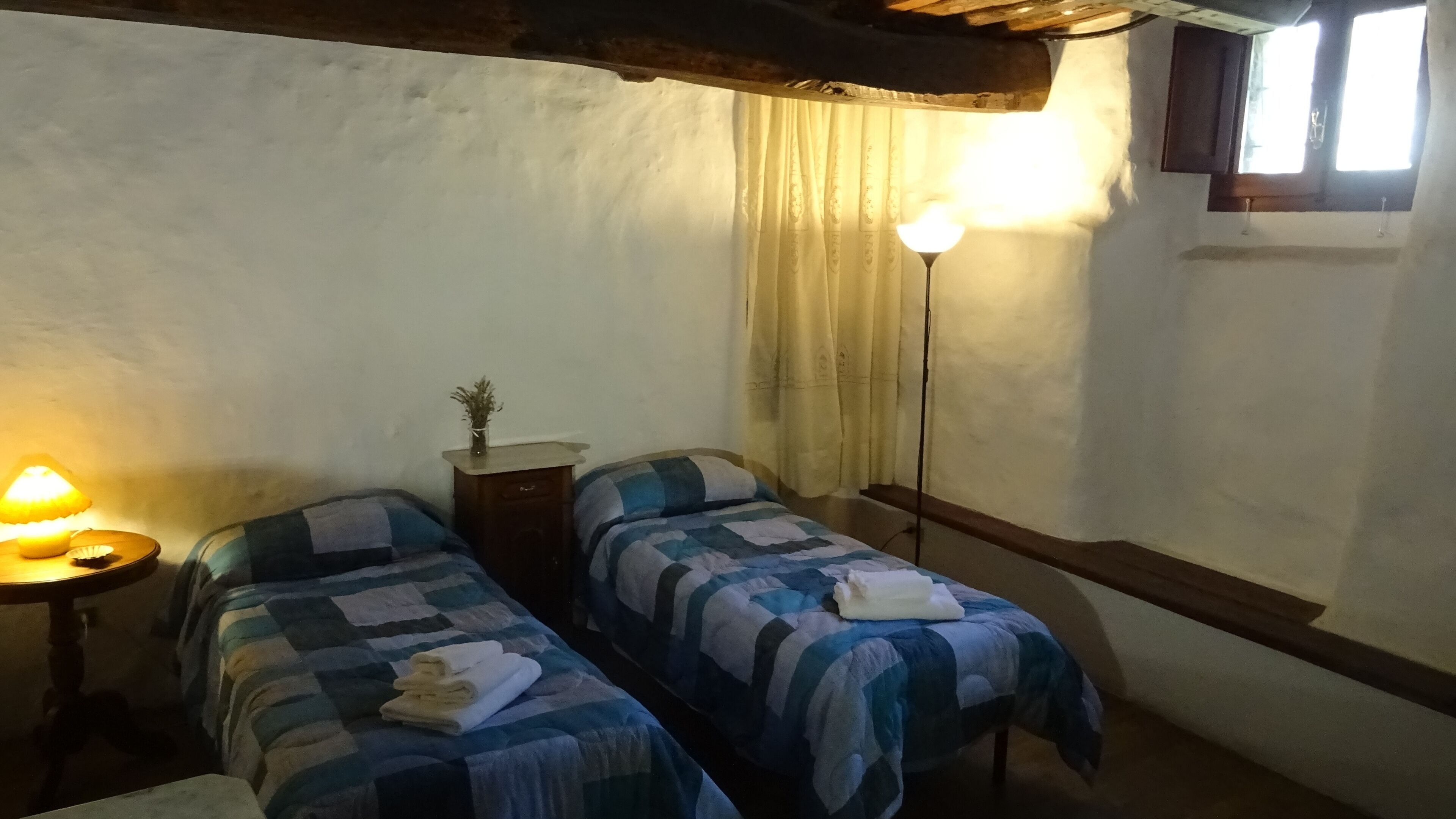 Een kleine, gezellige kamer met twee eenpersoonsbedden, een nachtkastje met een lamp, een tafeltje met een vaas, en een raam met gordijnen.