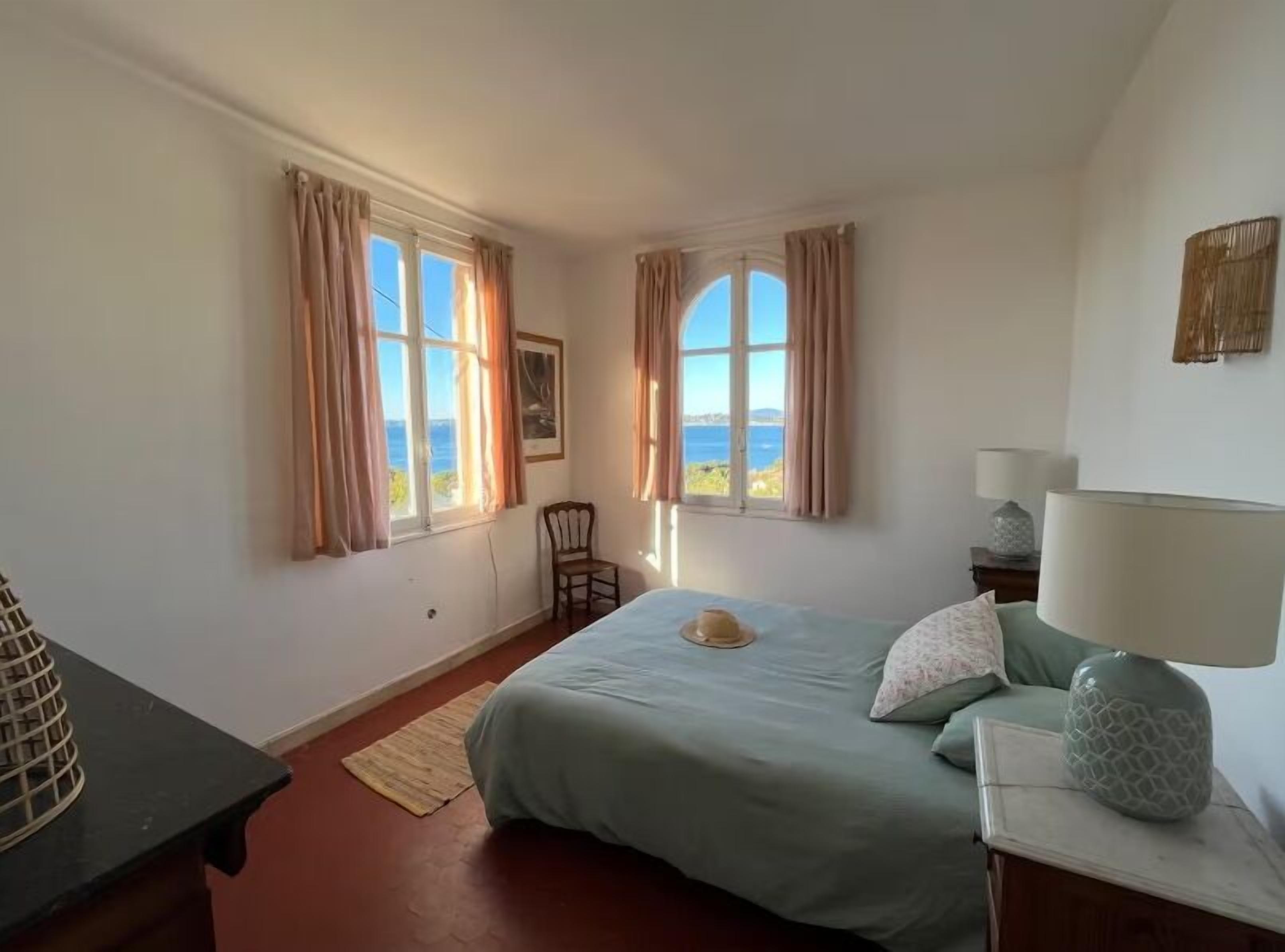 A bedroom with a bed, two windows with curtains, a chair, a lamp, and a view of the sea.