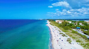 A coastal beach with clear turquoise waters, a sandy shore, and buildings along the coastline.