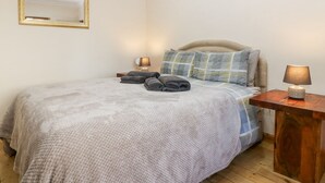 Una cama bien tendida con una manta gris y almohadas, una mesita de noche con una lámpara y un espejo en la pared.