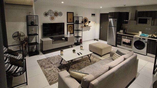 A modern living room with a sofa, TV, and a kitchen area with appliances. - Ebs properties  (Accra)