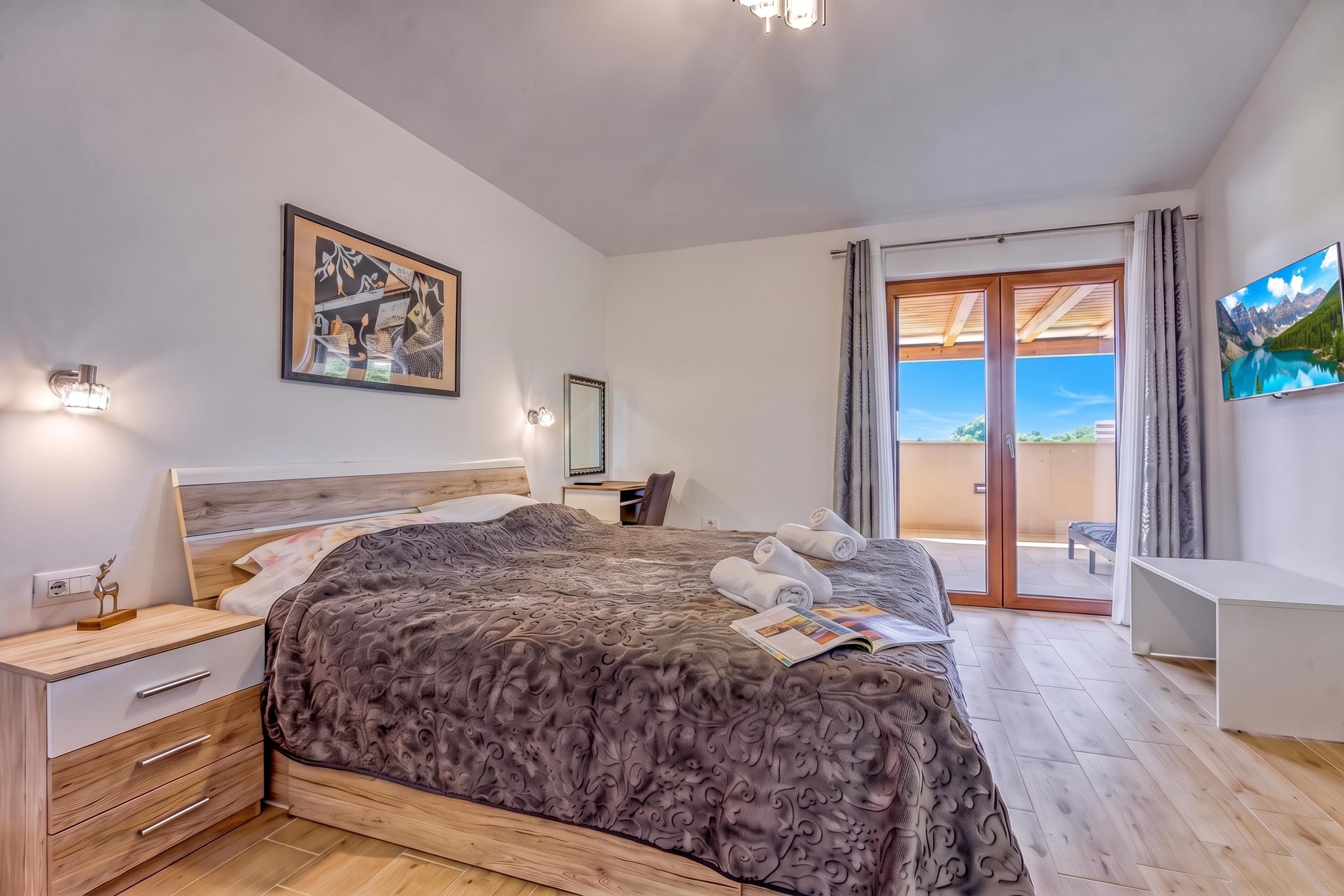 A bedroom with a bed, bedside tables, a desk, a chair, a painting, a mirror, and a view of the outdoors through a sliding door.