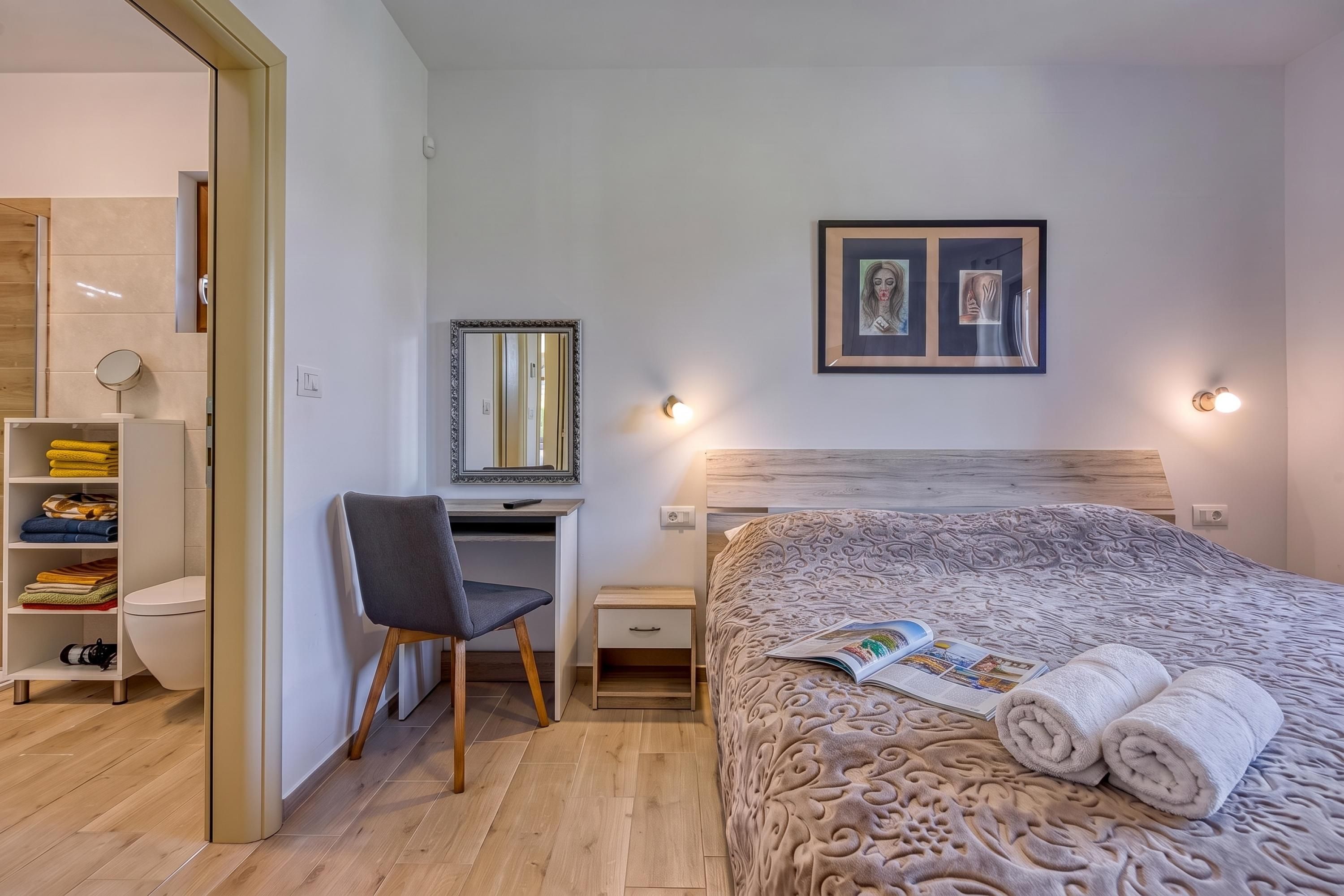 Quarto moderno com cabeceira de madeira, escrivaninha com cadeira, cama grande com colcha estampada e espelho fixado na parede.