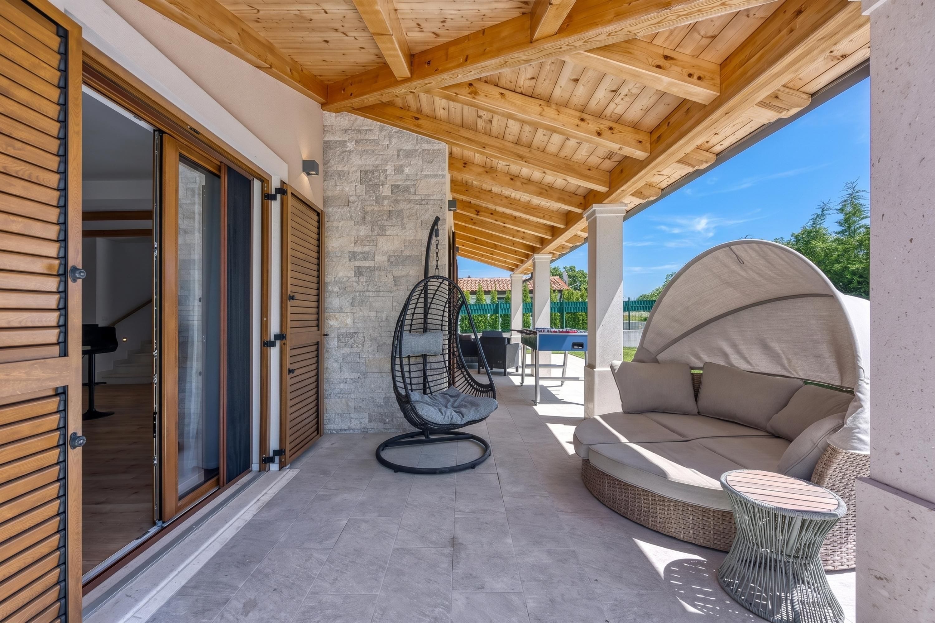 A covered outdoor patio with a wooden ceiling, a stone wall, a swing chair, a sofa, and a table.