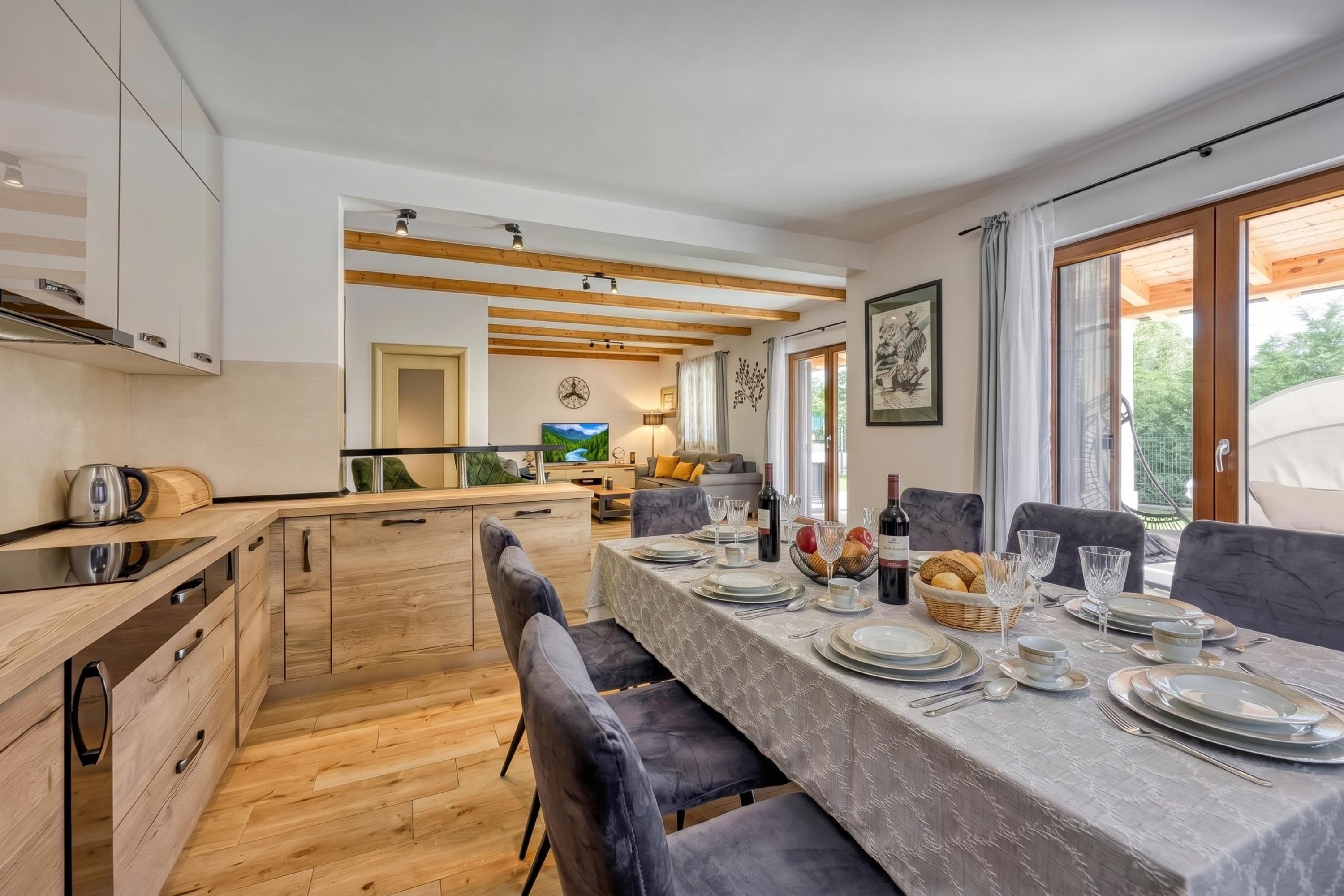 Un espace de restauration moderne avec une longue table dressée pour un repas, des meubles de rangement en bois et une vue sur l’extérieur.