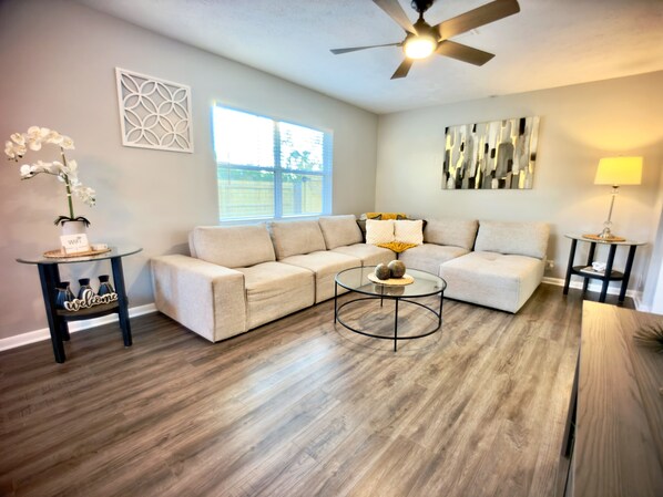 A living room with a sectional sofa, a glass coffee table, a ceiling fan, and a window with a view. - Heated Pool & Basketball, Sleeps 12 | Tropical Dream Villa, Valrico / Brandon (Valrico)