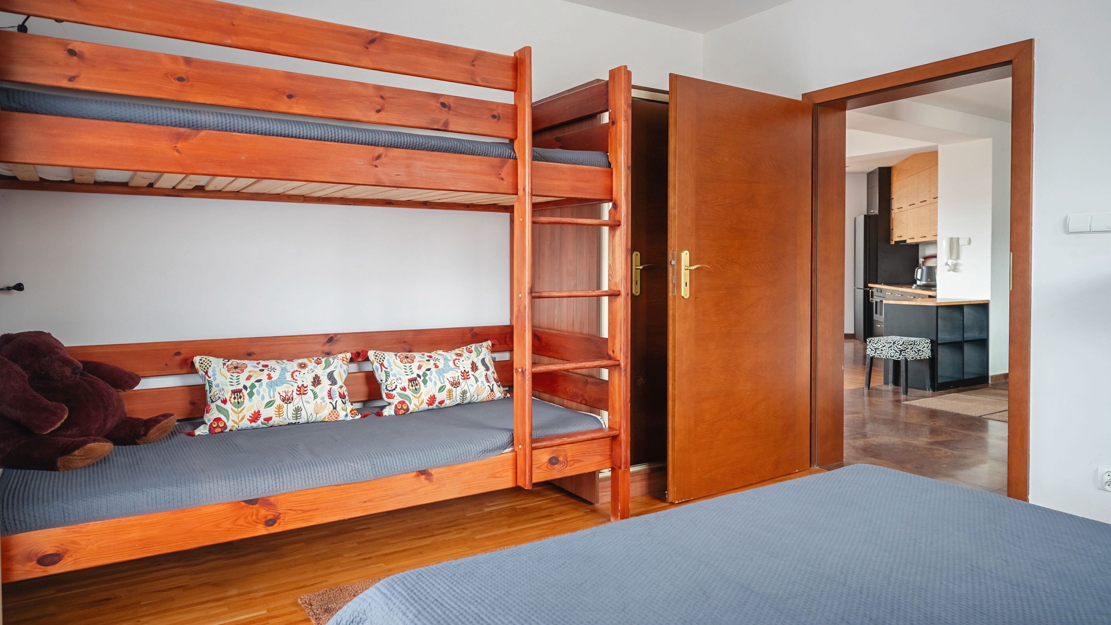 A bunk bed with a wooden frame and a single bed with a grey mattress. There is a teddy bear on the bunk bed and a patterned pillow. A wooden door is open, leading to a kitchen area.
