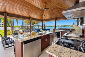 A covered outdoor kitchen with a view of the water, a dining area with chairs, and a pool in the background. - Stunning 5-Bedroom Lakefront Retreat! (Tampa)