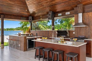 Une cuisine extérieure abritée, équipée d’un barbecue, de tabourets de bar et offrant une vue sur l’eau.