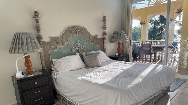 Un dormitorio con una cama, dos mesitas de noche con lámparas, una mesita auxiliar, un tapete estampado y vistas a un espacio exterior con sillas y una mesa.