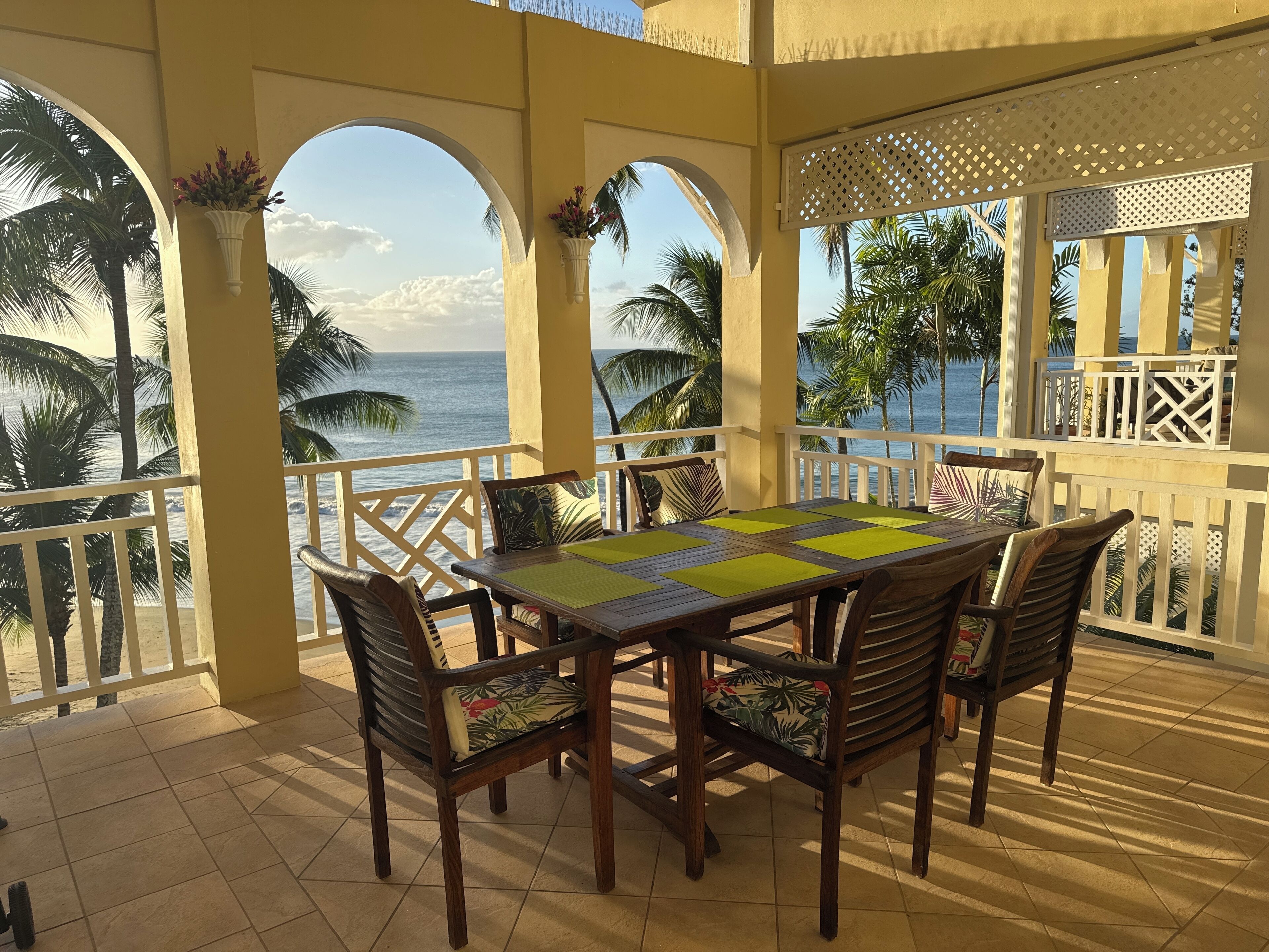 A covered outdoor dining area with a view of the ocean, featuring a wooden table and chairs, and a white railing.