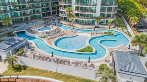 A multi-story hotel with a large outdoor swimming pool area, including a central pool and smaller pools, surrounded by lounge chairs and palm trees. - Villa Grande (Orange Beach)