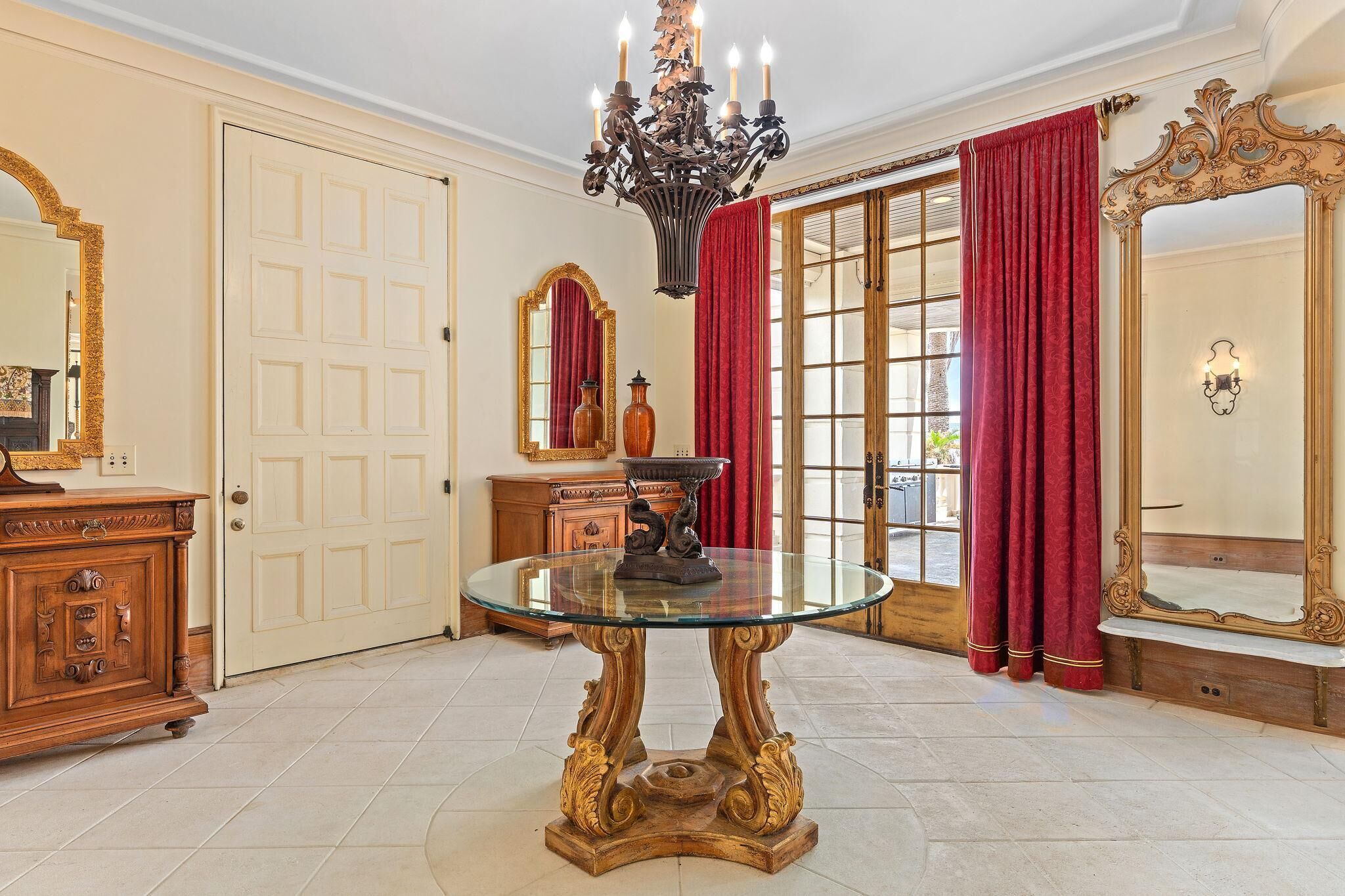 A room with a glass-top table, ornate wooden furniture, red curtains, and a chandelier.