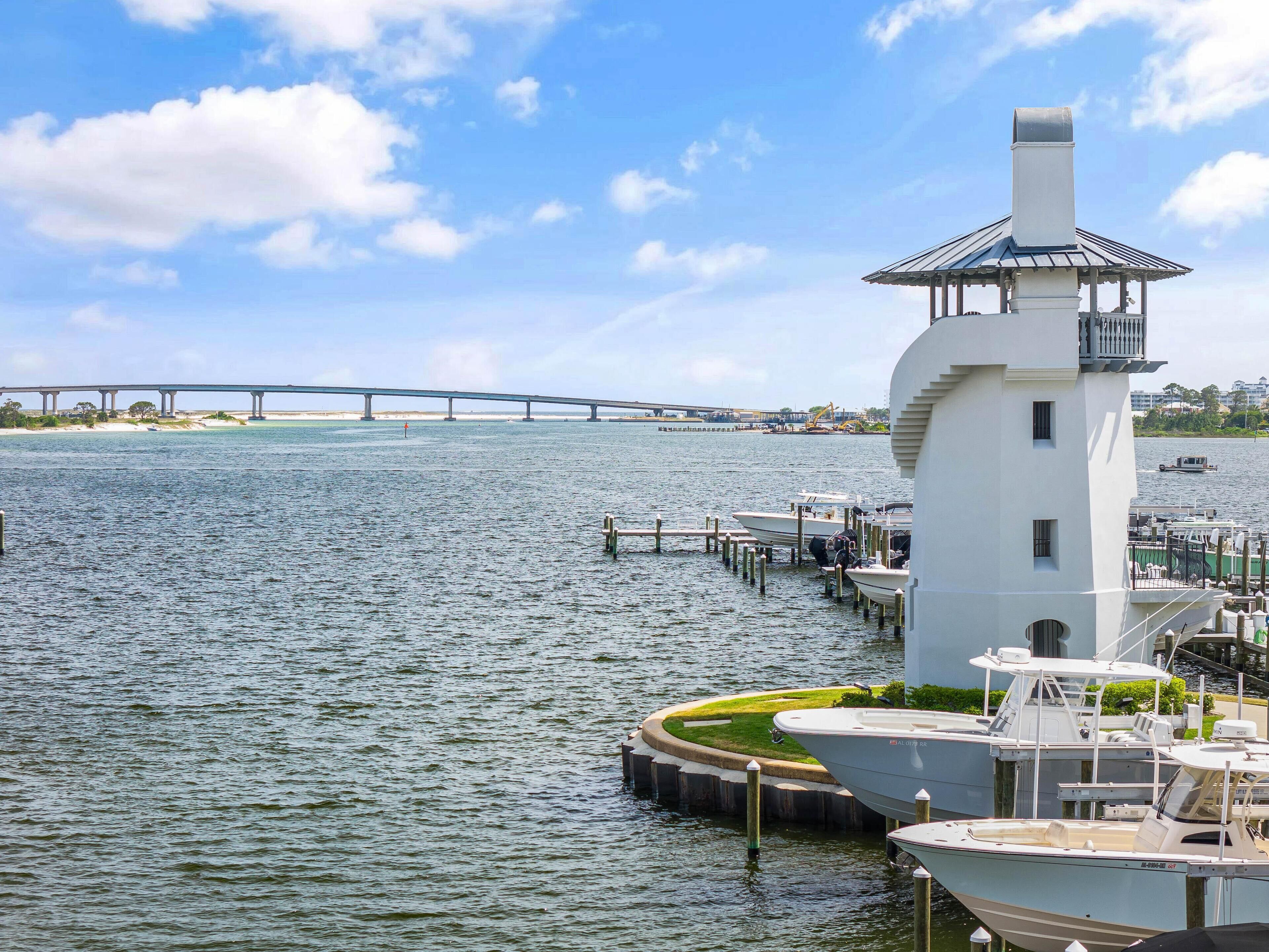 Un faro de exterior blanco con un balcón, barcos amarrados y un puente al fondo.