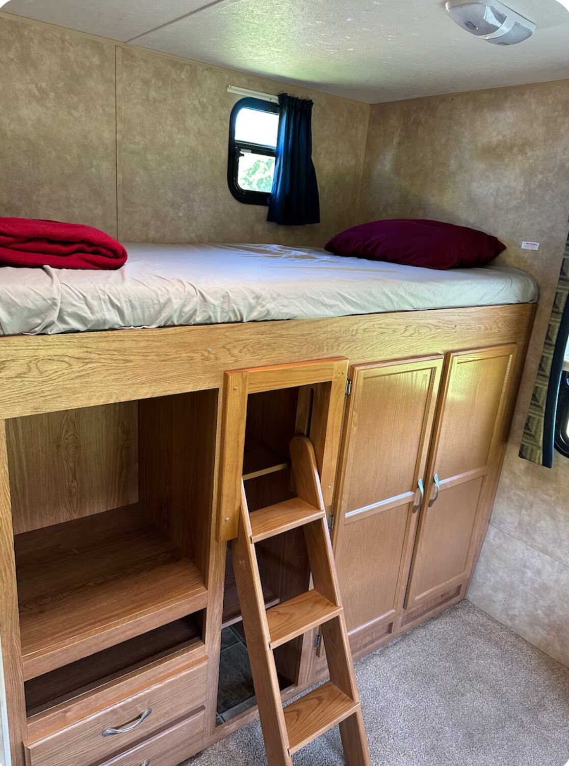A compact RV bedroom with a bunk bed, a small window with curtains, and a wooden ladder leading to a lower bunk.
