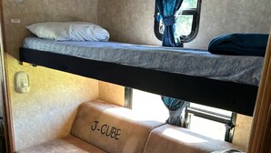 Een compact camper interieur met een bunkebed, een bank met de tekst 'J-CUBE' en een klein raam met gordijnen.