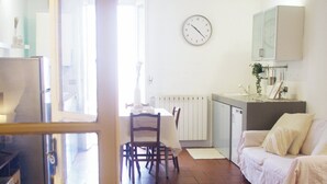 Een kamer met betegelde vloer, een eettafel met stoelen, een bank, een keukenhoek met een spoelbak en kasten, en een wandklok.