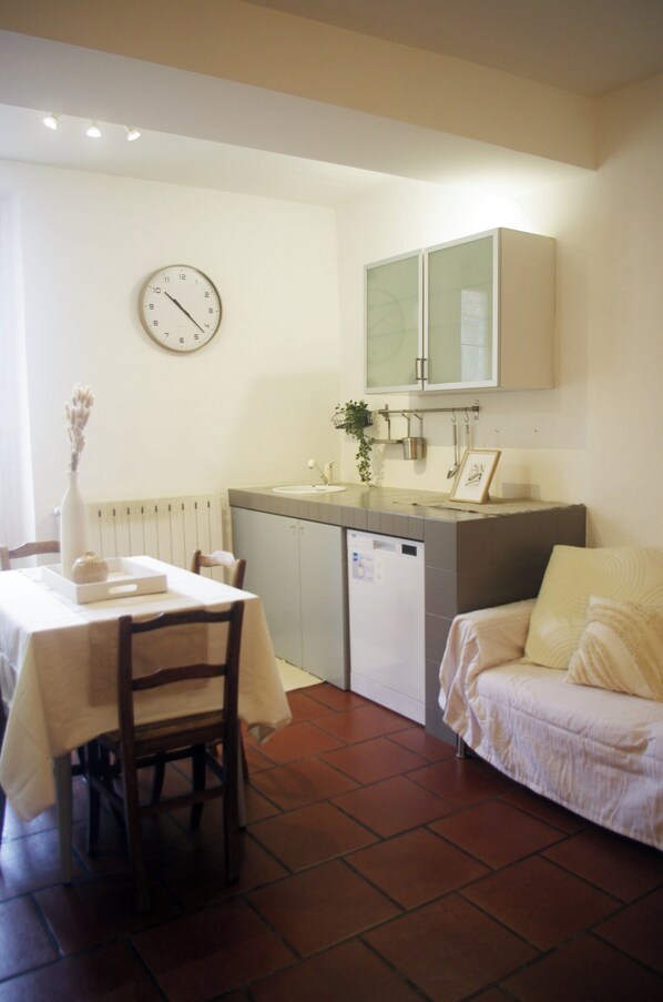 A compact kitchen with a dining table, chairs, a clock, a cabinet, and a small bed.