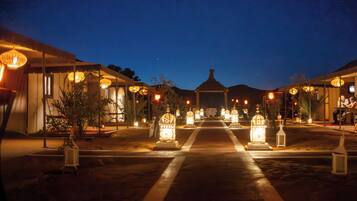 Acampamento no deserto com um caminho central que leva a um gazebo, ladeado por lanternas iluminadas e áreas de estar ao ar livre.