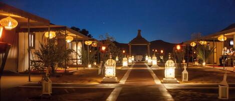 A desert camp with a central pathway leading to a gazebo, flanked by illuminated lanterns and outdoor seating areas.