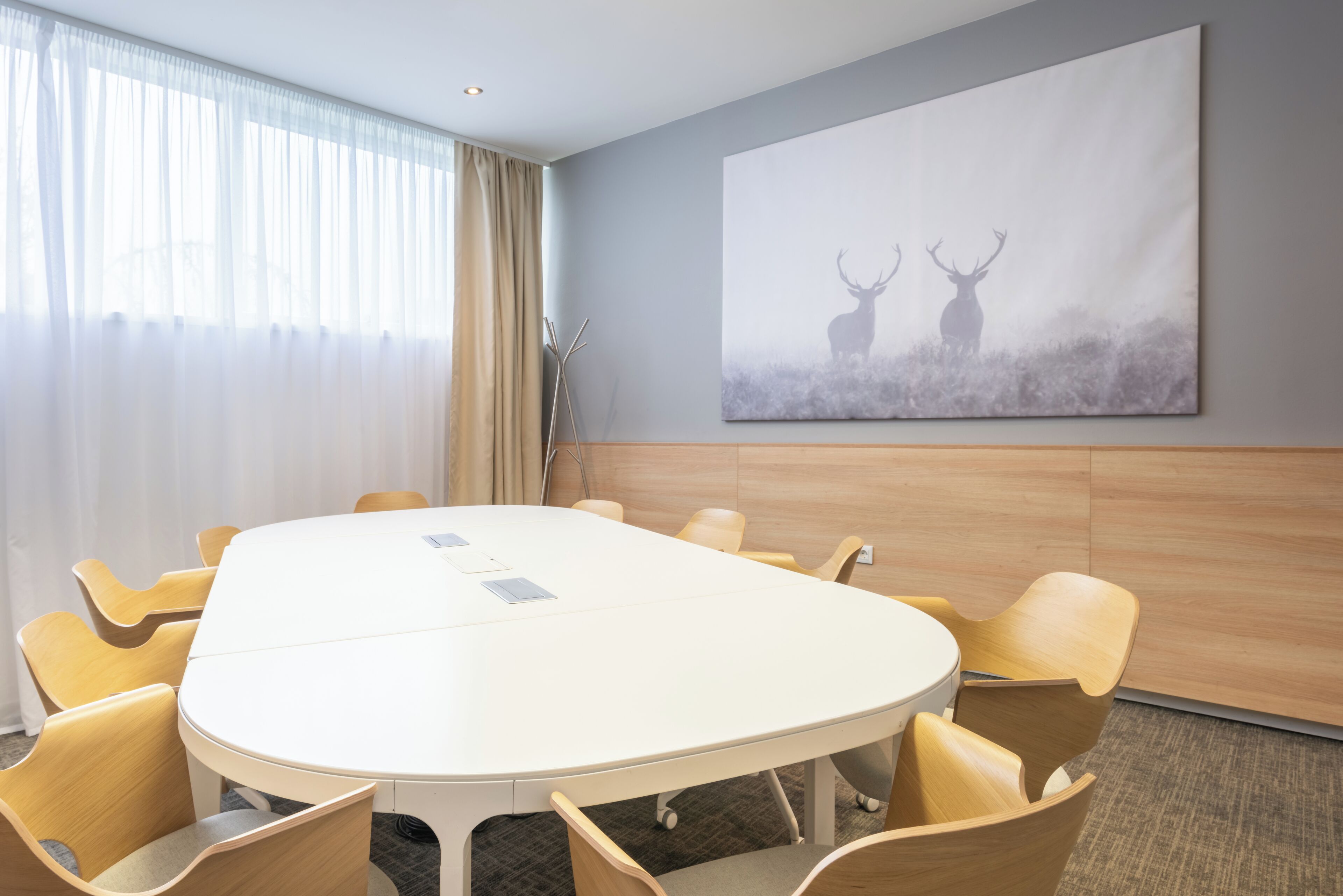 Un salón de conferencias con una mesa blanca, sillas color beige, un cuadro grande de ciervos y una ventana con cortinas traslúcidas.