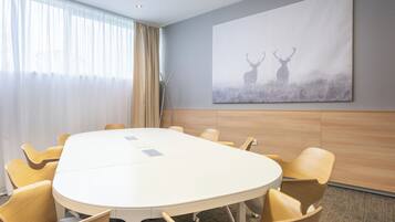 Un salón de conferencias con una mesa blanca, sillas color beige, un cuadro grande de ciervos y una ventana con cortinas traslúcidas.